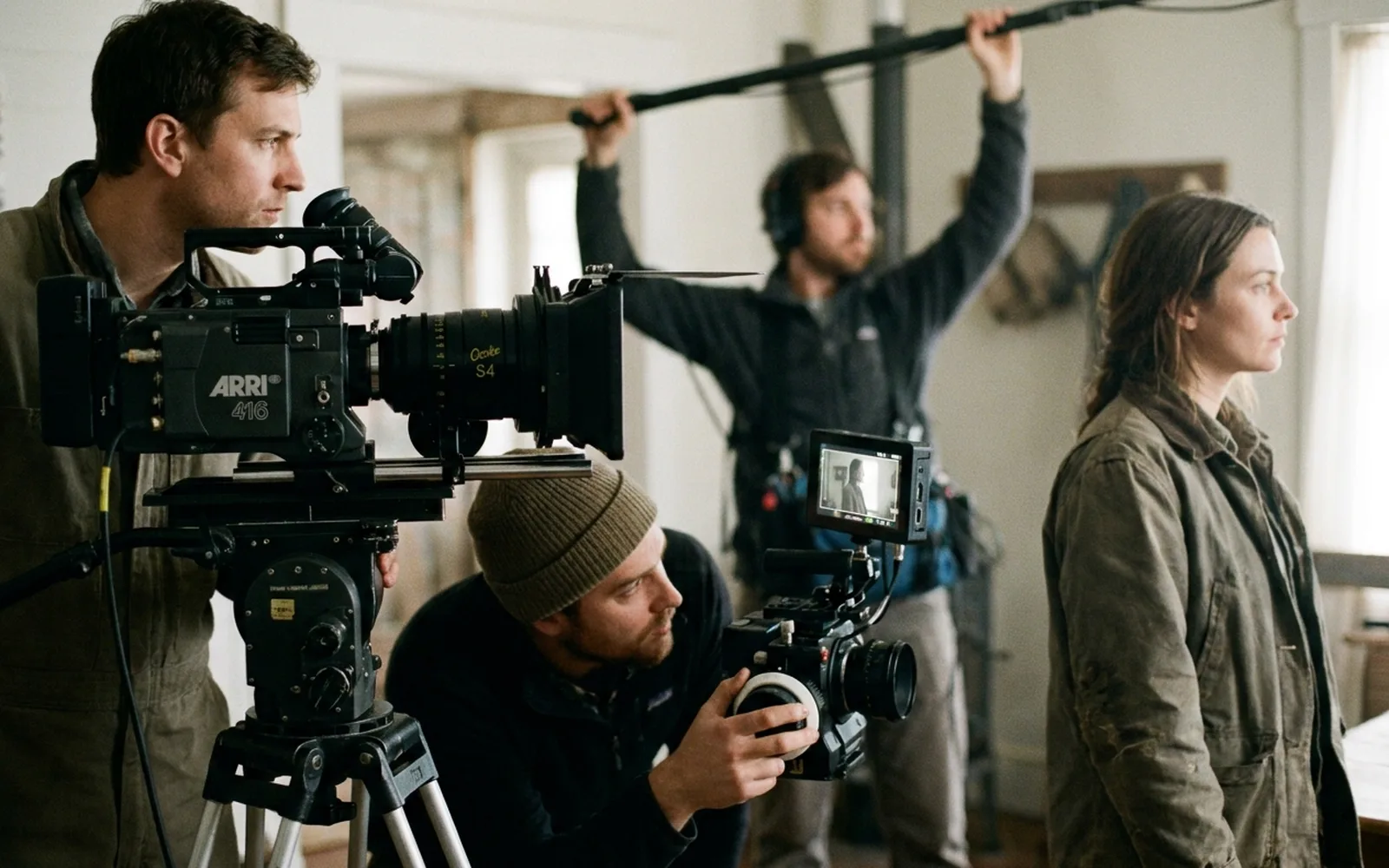A film crew shooting a scene indoors, with a camera operator at a tripod-mounted cine camera, a focus puller at a second camera and a boom operator holding a microphone overhead.