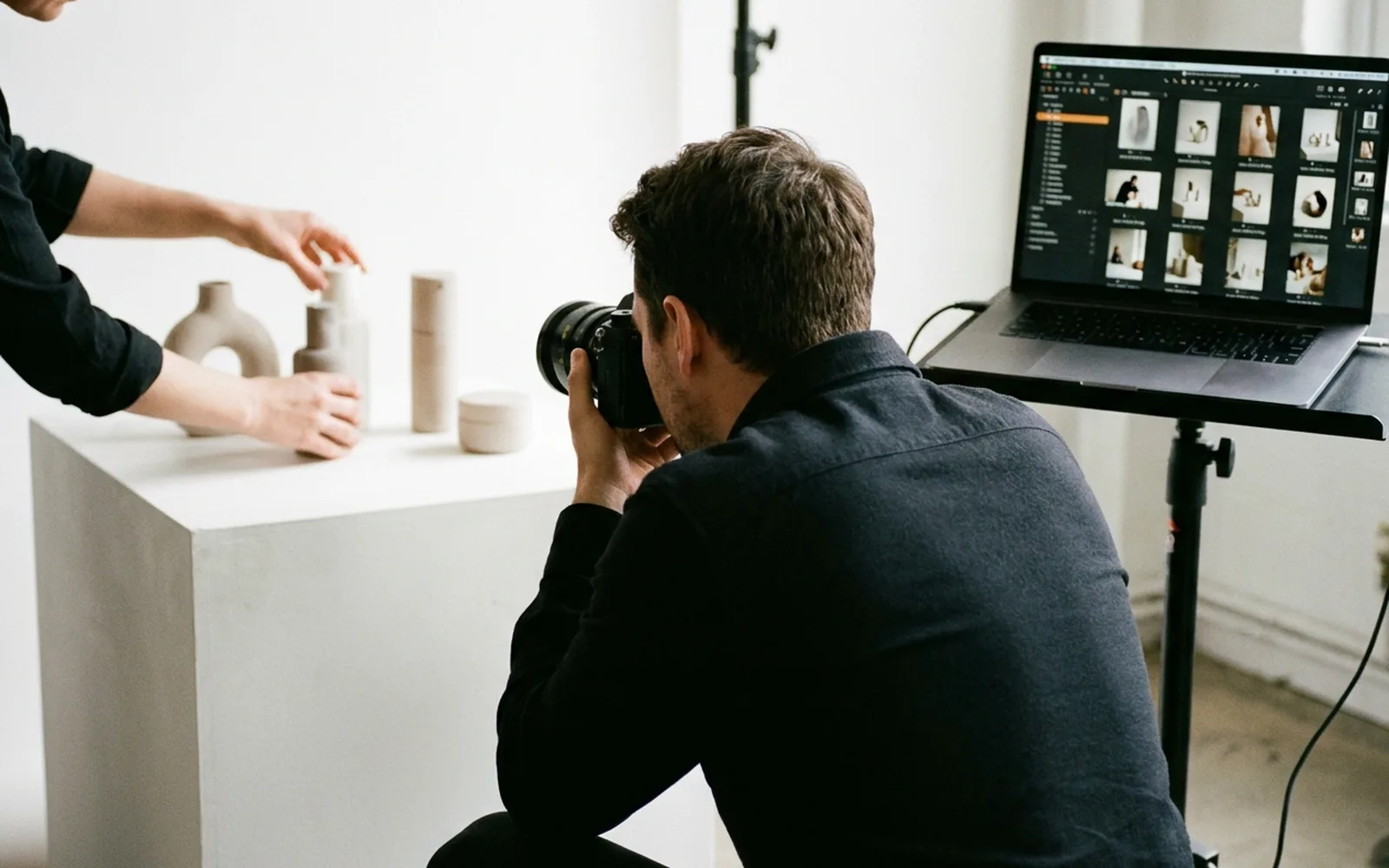 A photographer shooting a still-life on a white plinth while a stylist adjusts the products, with a tethered laptop showing the captures.