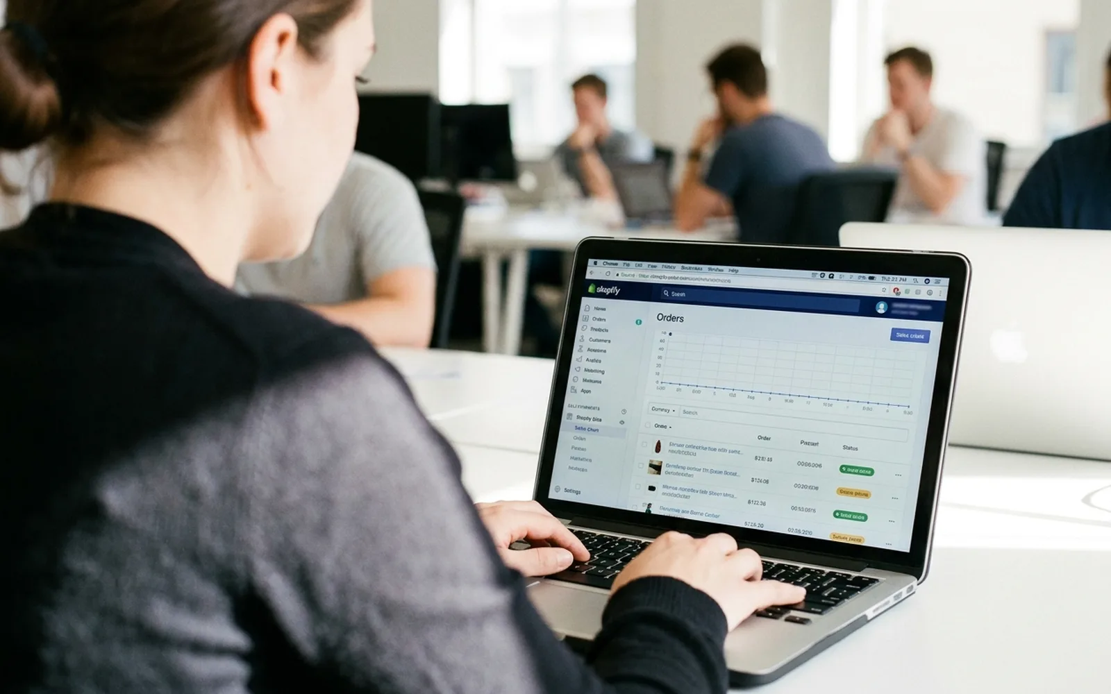 A woman working at a laptop in a bright open-plan office, an e-commerce orders dashboard open on screen.
