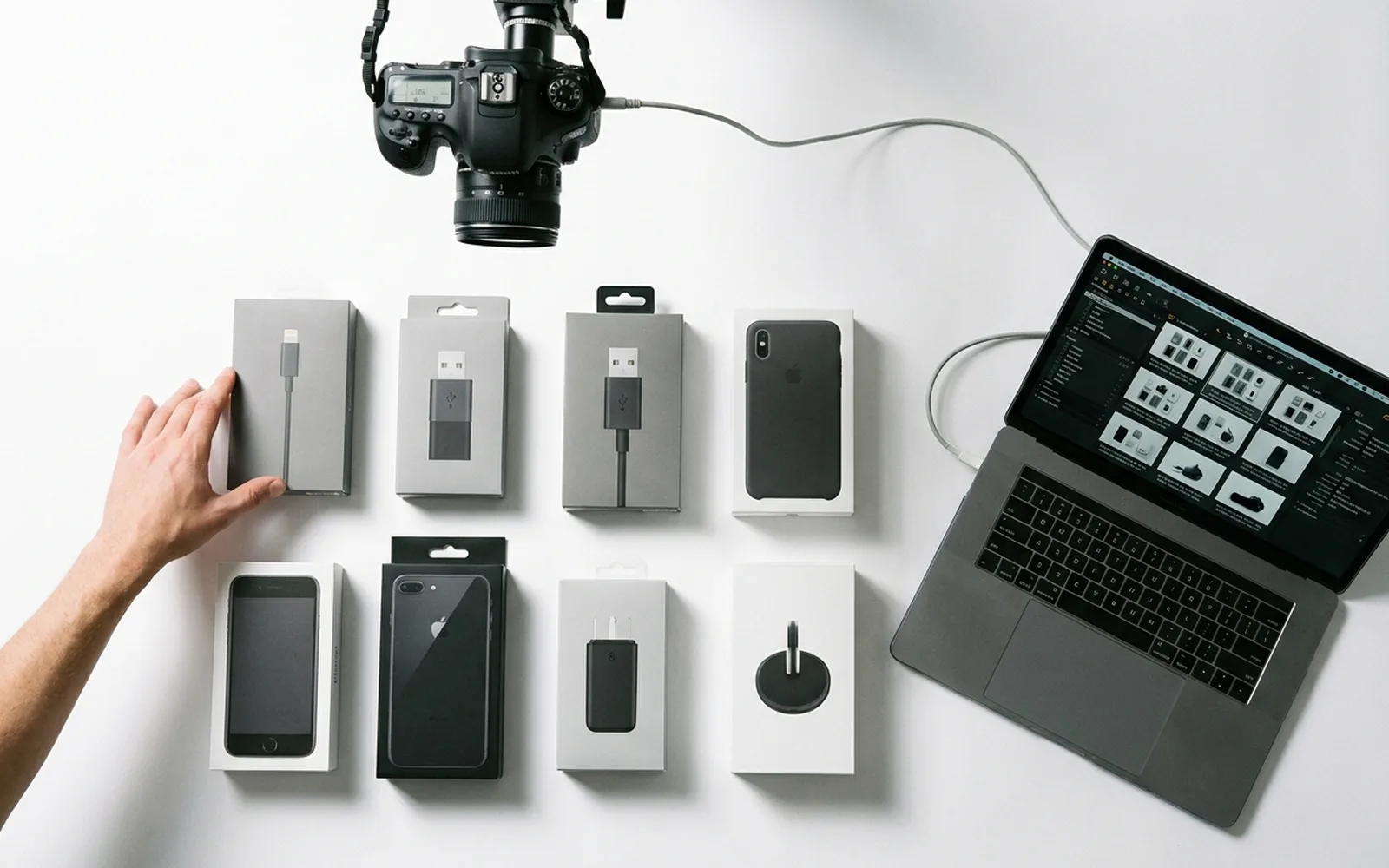 Boxed electronics accessories arranged on a white surface beside a camera on a copy stand and a laptop displaying the photographed product images.