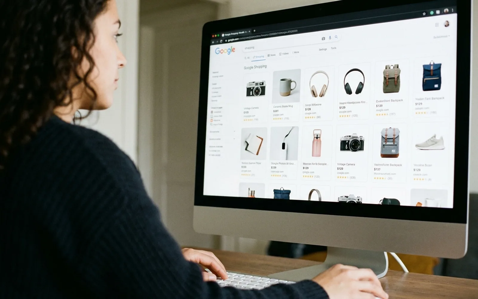 A woman at a desktop computer viewing a Google Shopping results page filled with product listings.
