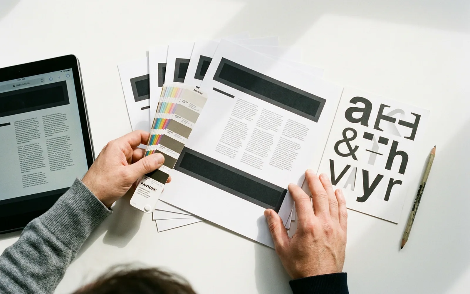 An overhead view of printed page layouts, a colour fan and a type specimen sheet on a white desk, with a tablet showing the same layout.