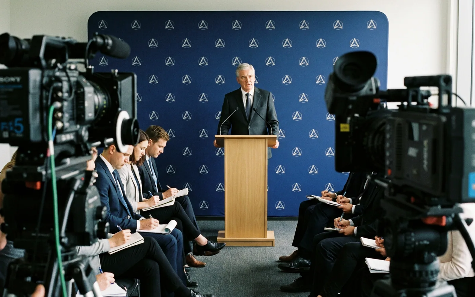 A speaker at a lectern with two microphones, seen from the back of a press room past several broadcast cameras on tripods and a seated audience.