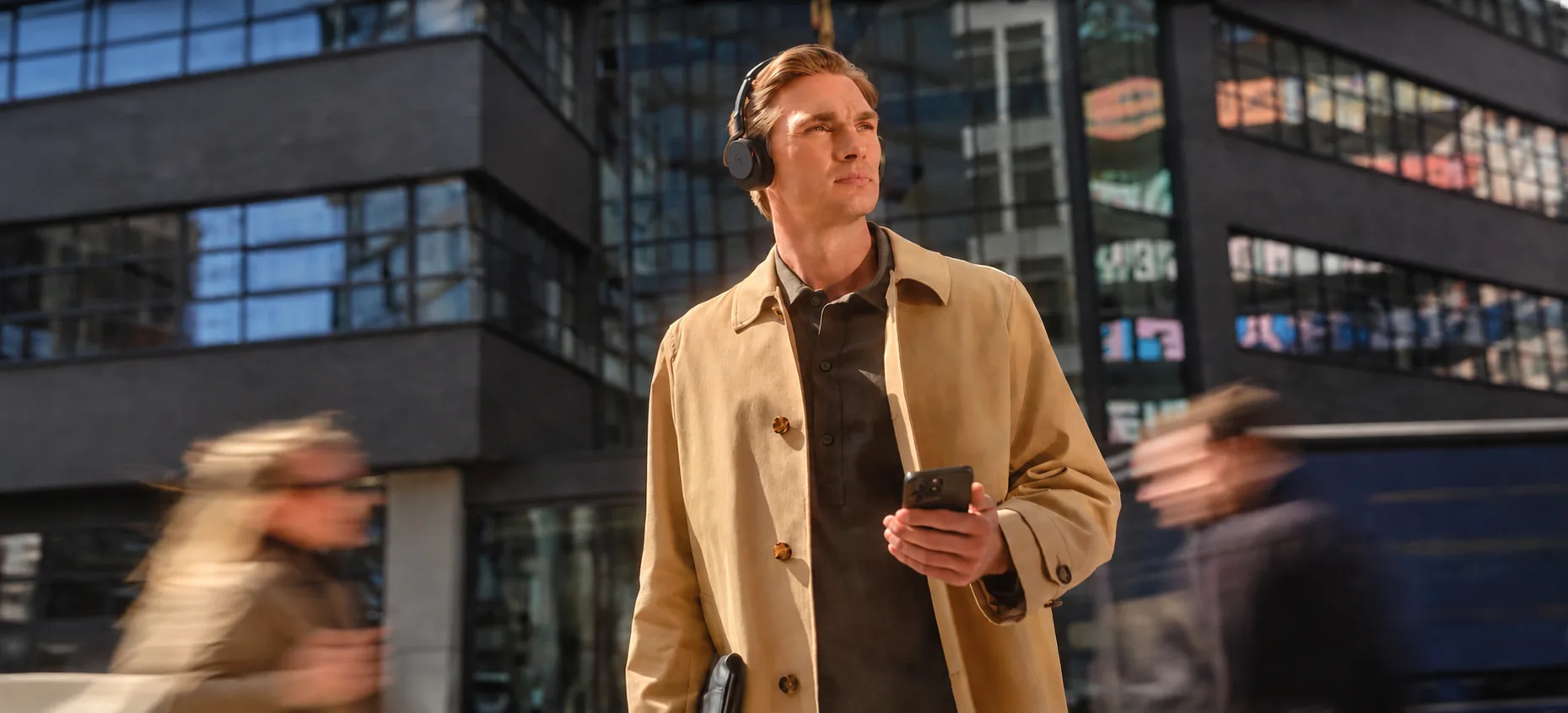 A man wearing over-ear headphones walking through a city street, checking his phone