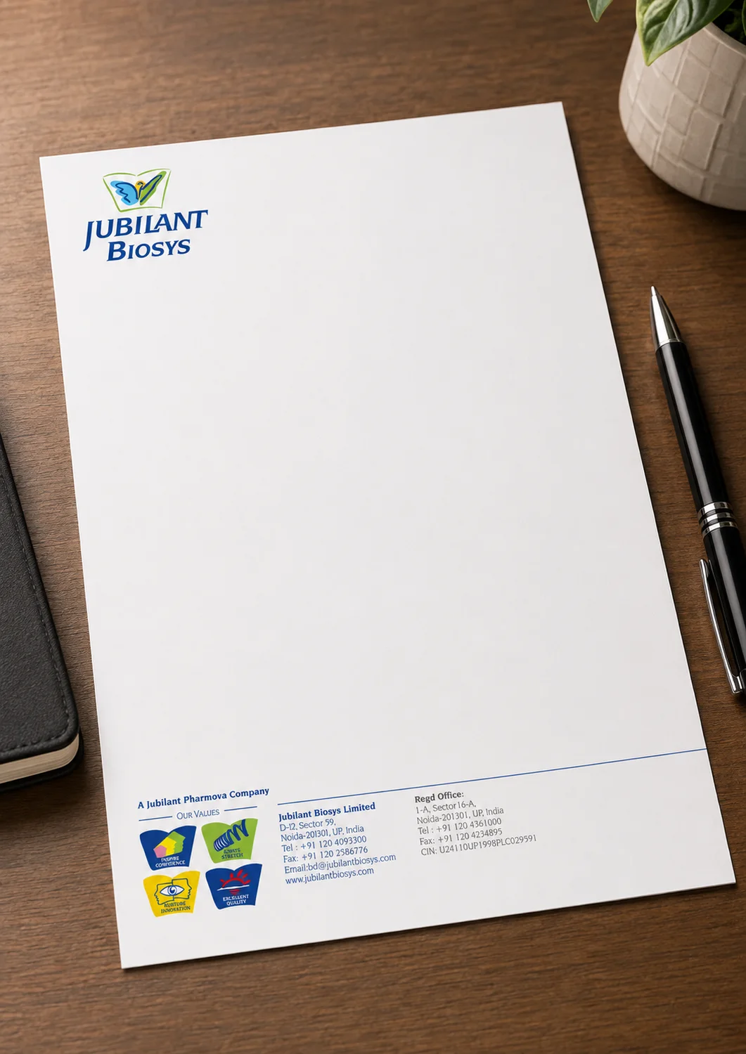 Jubilant Biosys letterhead on a wooden desk beside a pen and notebook, showing the logo, the four company values marks and the registered office details